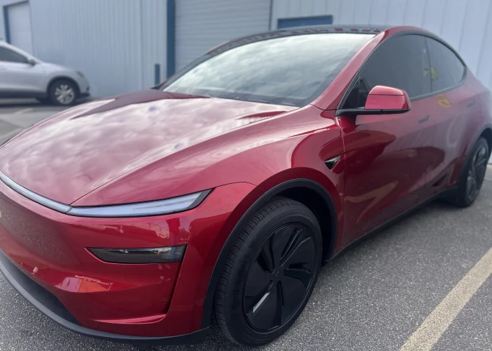 Sleek red Tesla Model Y with dark tinted windows and black aerodynamic wheels parked in front of a commercial building. The car's glossy finish reflects a cloudy sky.