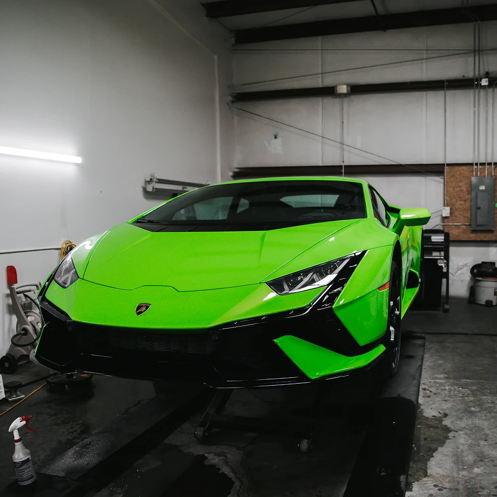 Bright green Lamborghini sports car positioned on a lift inside a detailing garage, with a bottle of cleaning solution visible on the floor nearby.