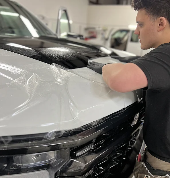 Technician applies paint protection film to the hood of a white truck, using a squeegee to smooth the film over wet surface.