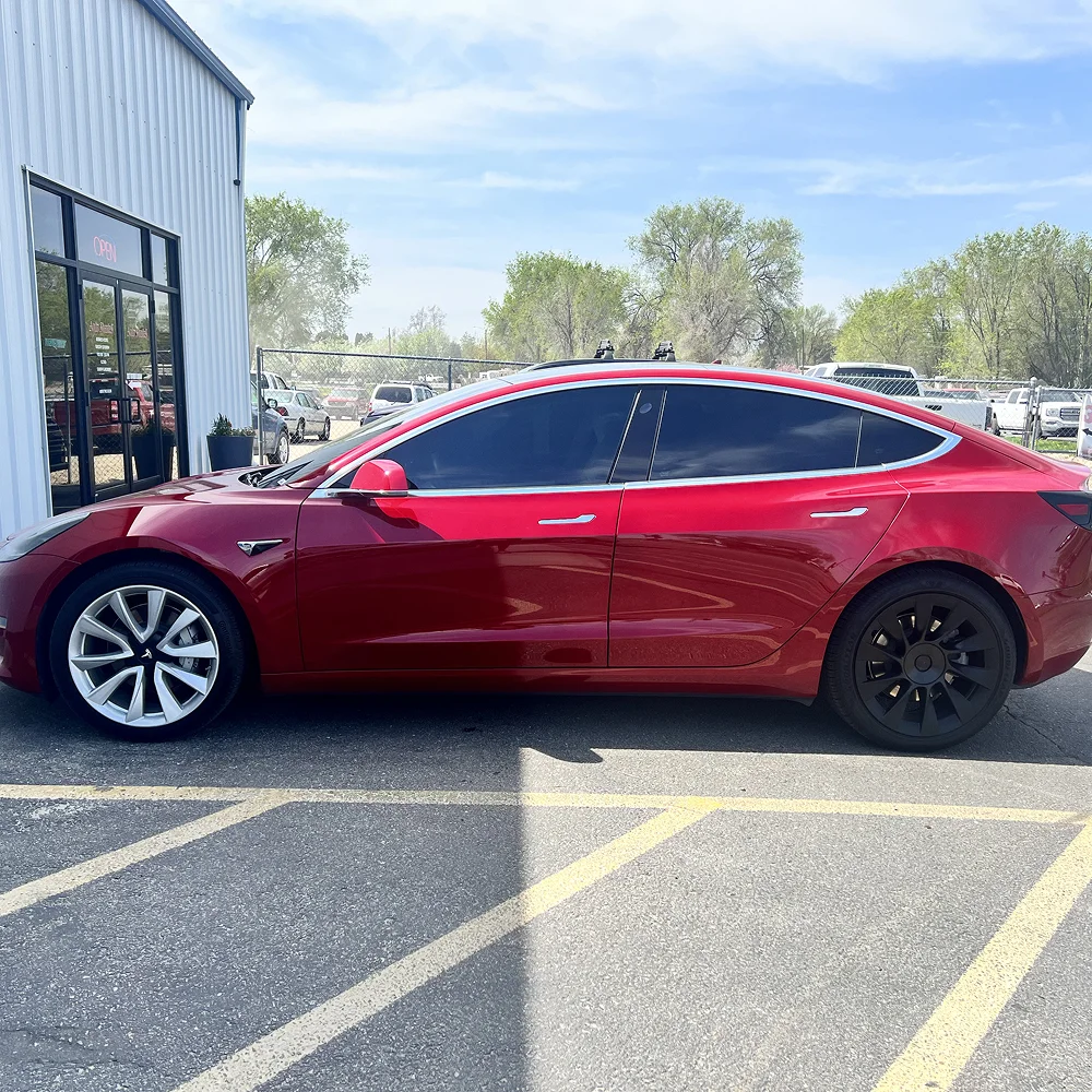 Shiny red Tesla Model 3 with dark tinted windows parked outside a building, featuring mismatched wheels—silver multi-spoke on the front and matte black aero-style on the rear.