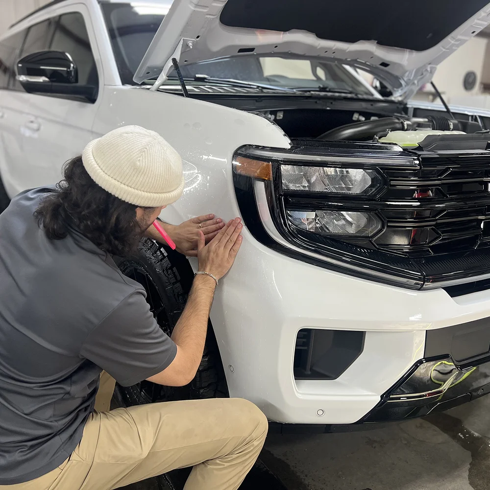 Technician wearing a white beanie and gray shirt applies clear protective film to the front fender of a white SUV with the hood open inside a detailing shop.