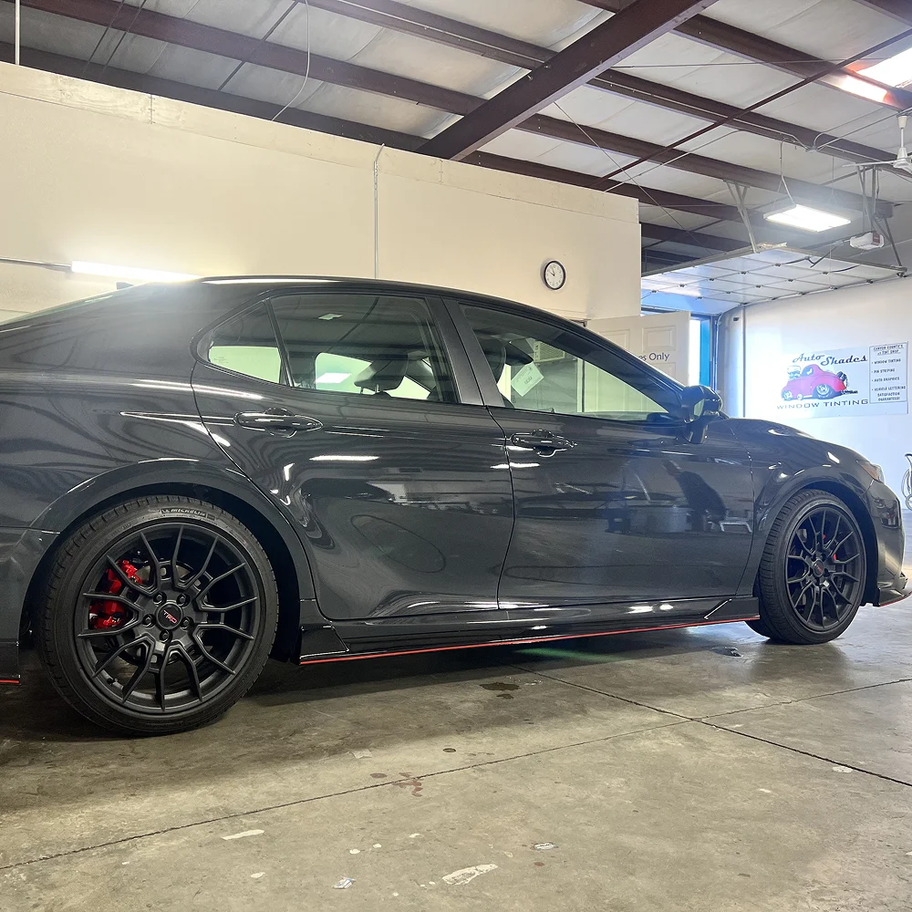 Glossy black sedan with red brake calipers and black multi-spoke wheels parked inside an auto shop with a sign in the background reading "Auto Shades Window Tinting."