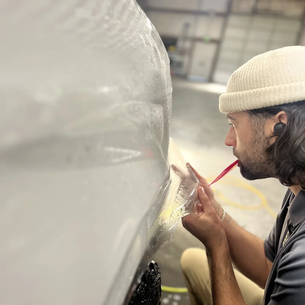Close-up of a technician in a white beanie applying clear protective film to a white vehicle bumper inside a workshop, holding a red squeegee tool between his lips.