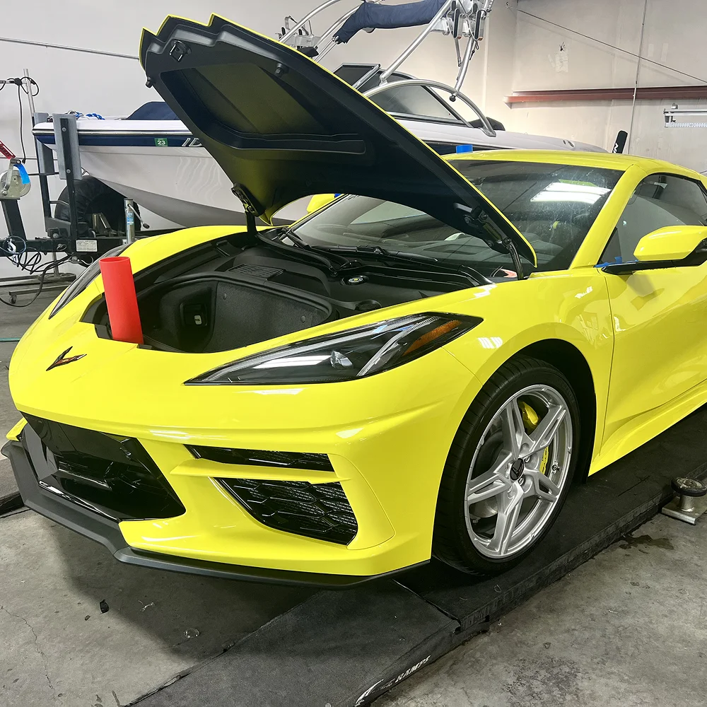 Bright yellow Chevrolet Corvette with the front trunk open, parked inside a garage next to a boat, with a red cylinder placed inside the frunk.