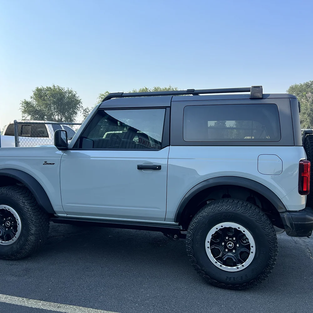 Light blue two-door Ford Bronco with black fender flares and off-road tires parked on asphalt near a chain-link fence and other vehicles, under a clear blue sky.