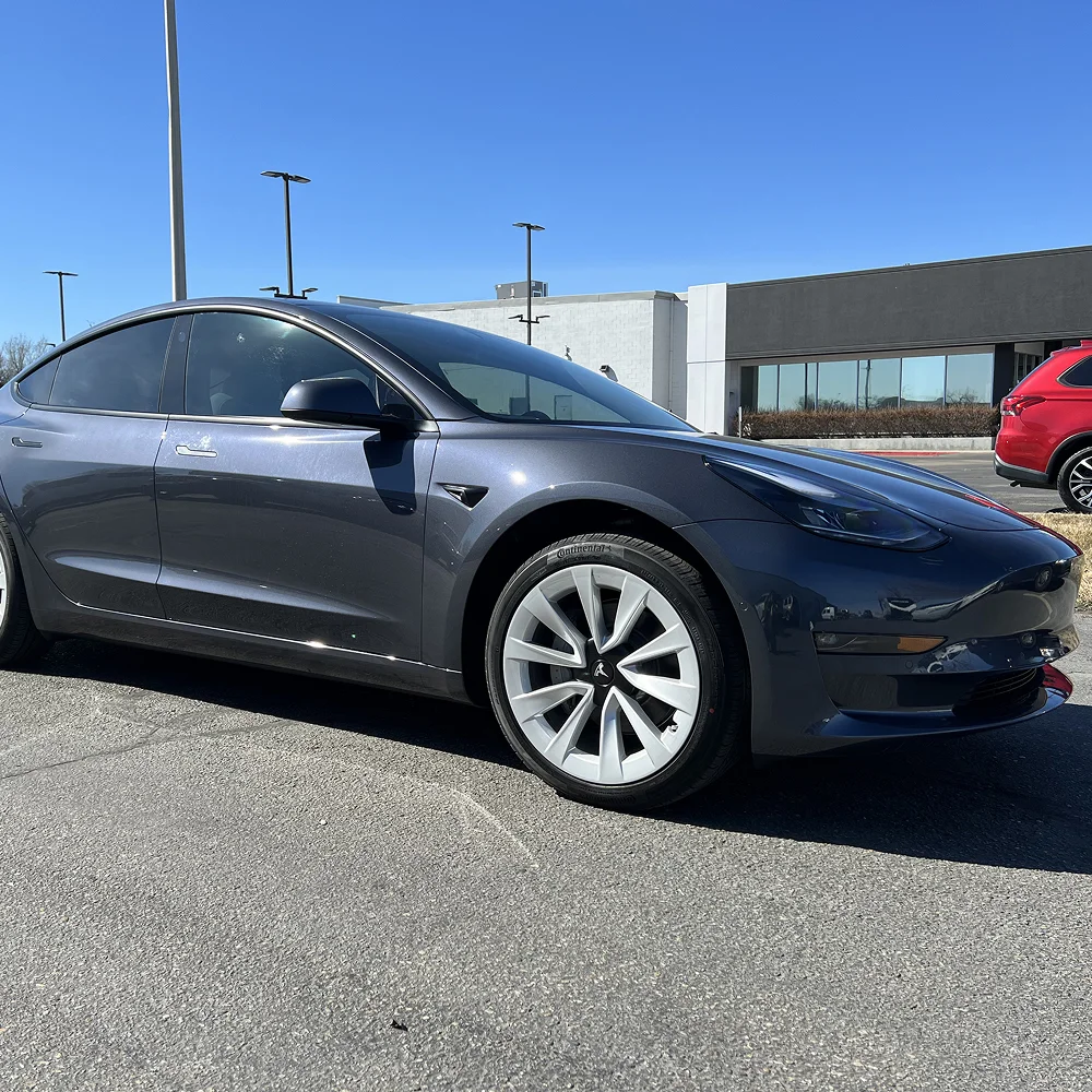 Glossy dark gray Tesla Model 3 with silver aero wheels parked in a sunny lot near a modern dealership building under a clear blue sky.