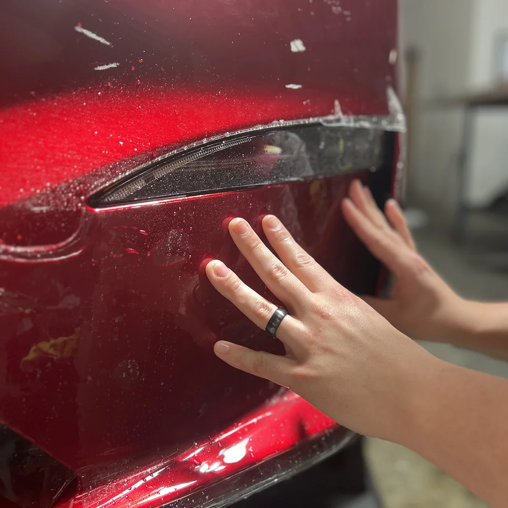 Close-up of hands applying a clear protective film over the headlight and bumper of a shiny red vehicle, with water droplets visible during the installation process.