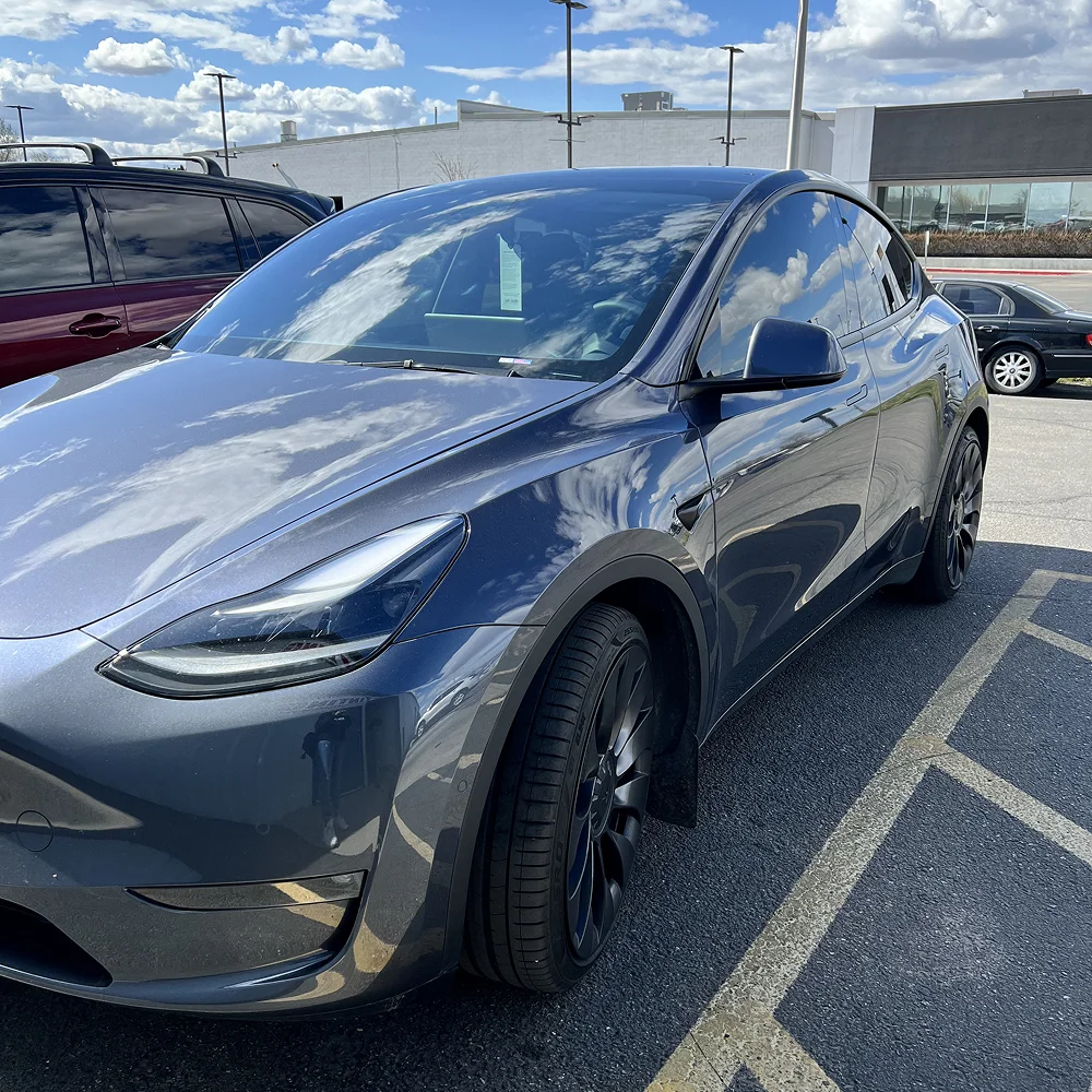 Dark gray Tesla Model Y with tinted windows and black wheels parked in a lot under a partly cloudy sky, with clouds vividly reflected on the vehicle's glossy surface.