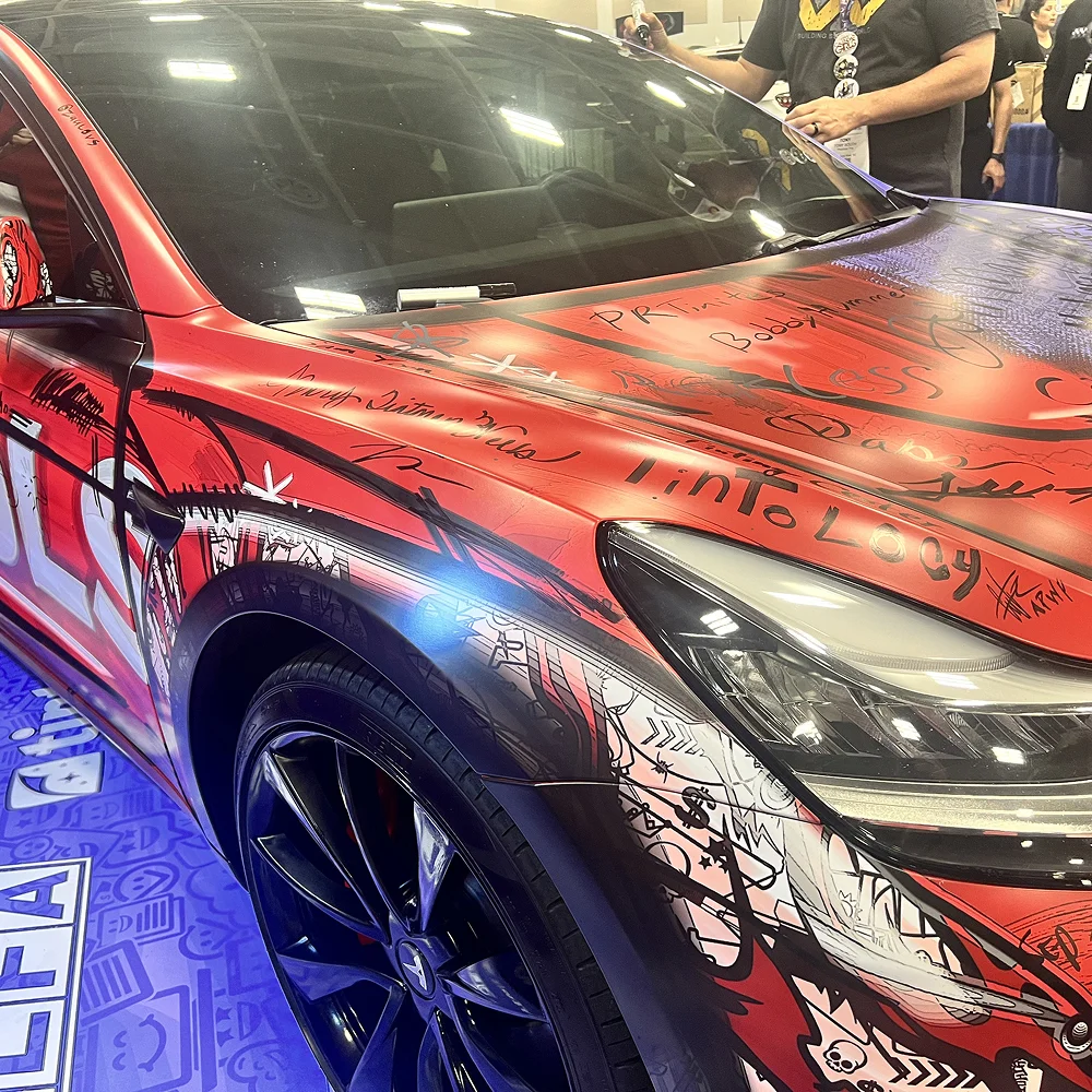 Custom-wrapped Tesla with a red, black, and white comic-style design covered in handwritten signatures, displayed indoors on a purple floor mat at an event with people gathered around.