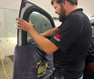 A technician carefully applies window tint film to a car’s front side window, which is partially rolled down and protected by a towel. The work is being done indoors with tools clipped to the technician’s belt.