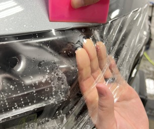 Close-up of a technician’s hands applying clear paint protection film to a vehicle bumper, using a pink squeegee to smooth out bubbles.