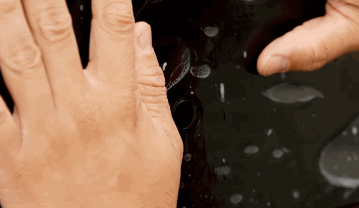 Hands smoothing out clear paint protection film on a glossy black car surface with water and air bubbles visible.