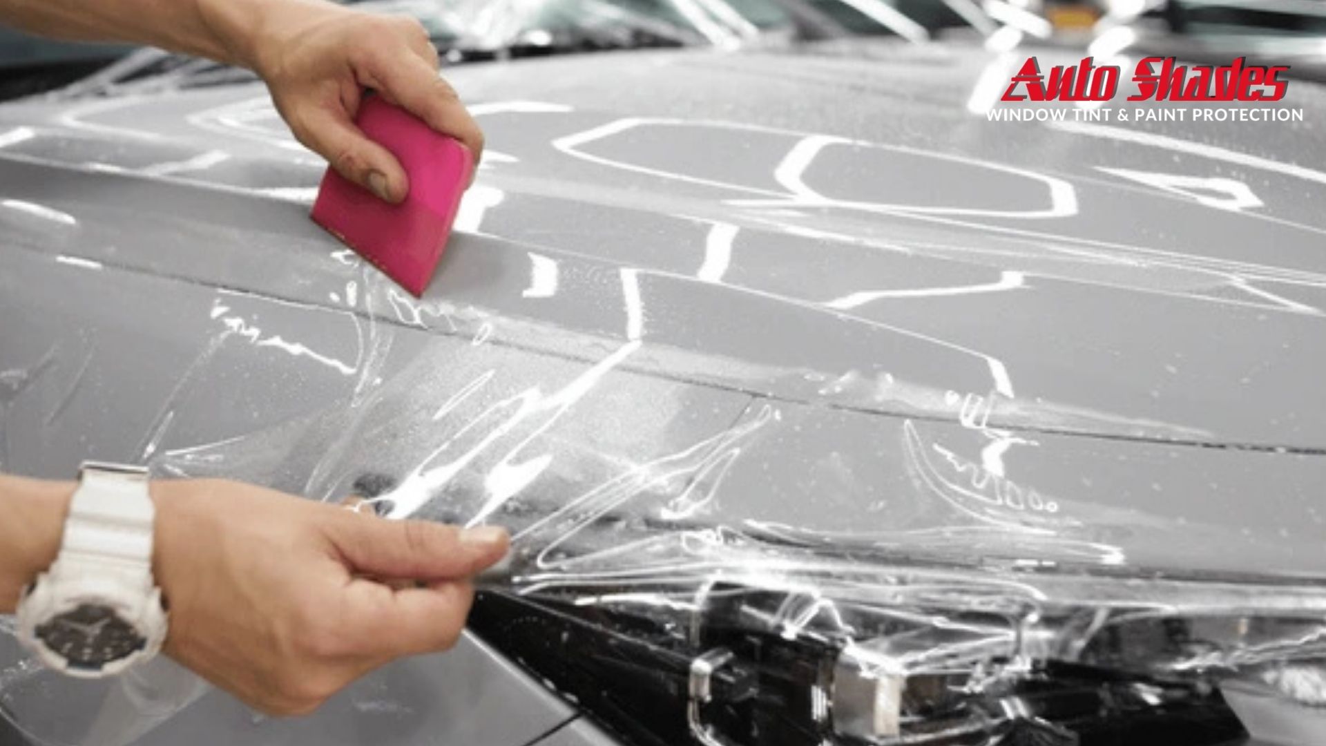 Hands applying a clear paint protection film to a car hood using a pink squeegee, with Auto Shades logo in the top right corner.