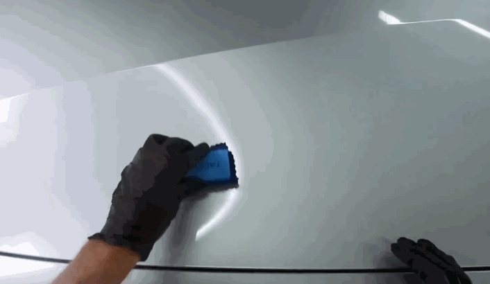 Close-up of a gloved hand spreading ceramic coating on a car’s white hood with a blue applicator pad, creating a high-gloss finish.