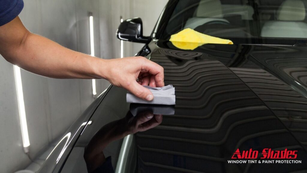 A hand using a folded microfiber cloth to apply ceramic coating on a glossy black car hood inside a detailing booth, with strong light reflections visible.