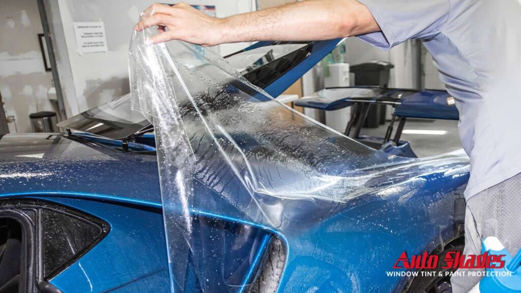 A worker carefully lays paint protection film over the rear of a blue sports car, with water droplets showing the installation process.