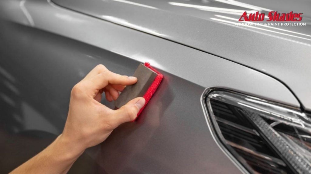 A hand uses a red microfiber applicator to spread ceramic coating across the surface of a silver car’s fender.