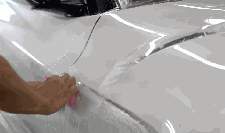 A hand uses a squeegee to smooth clear paint protection film over the curved fender of a white car during installation.