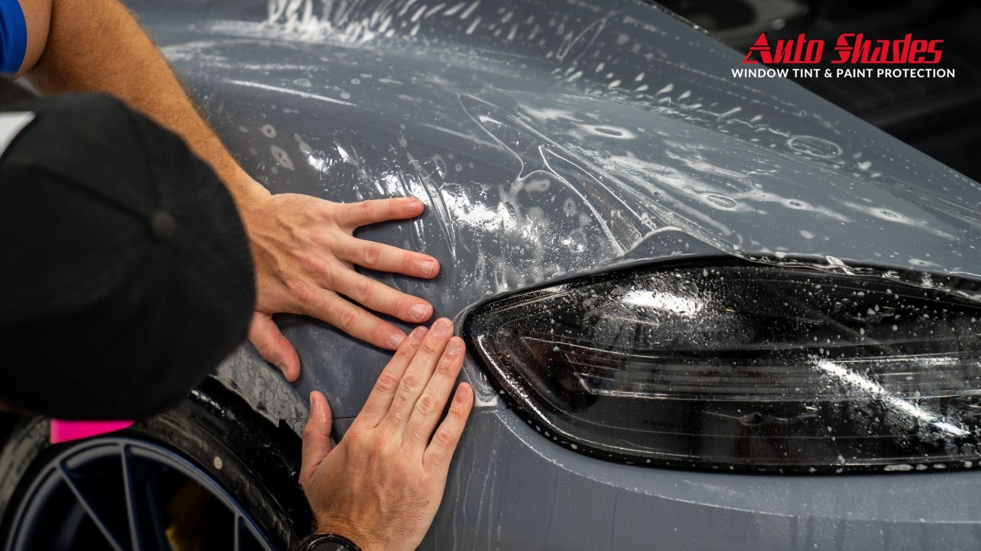 Installer presses paint protection film across the rear quarter panel and taillight of a gray car while water solution and bubbles sit beneath the clear film. The corner of the image shows the text "Auto Shades Window Tint & Paint Protection" as the paint protection film is carefully positioned and smoothed.