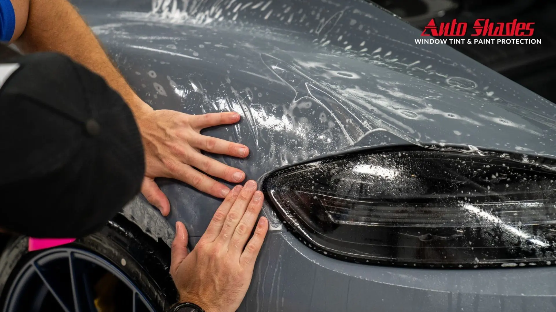 Installer presses paint protection film across the rear quarter panel and taillight of a gray car while water solution and bubbles sit beneath the clear film. The corner of the image shows the text "Auto Shades Window Tint & Paint Protection" as the paint protection film is carefully positioned and smoothed.
