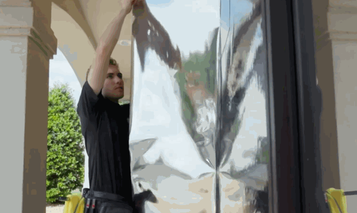 A worker holds a large sheet of commercial window tint against a tall glass panel outside a building. The reflective film is positioned before application showing how commercial window tint is measured and aligned.