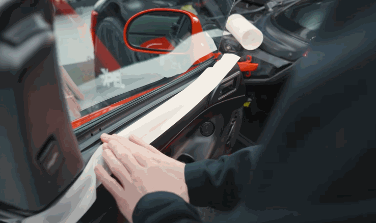 Technician inside a car carefully applies window tint film along the edge of a side window while smoothing it into place. The red exterior side mirror is visible, adding context to the precise window tint installation process.