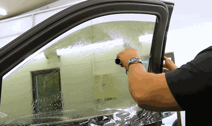 Installer uses a tool to smooth window tint film across a wet car window surface, removing bubbles during the application process. The glass is covered in water droplets to ensure a clean and even window tint finish.