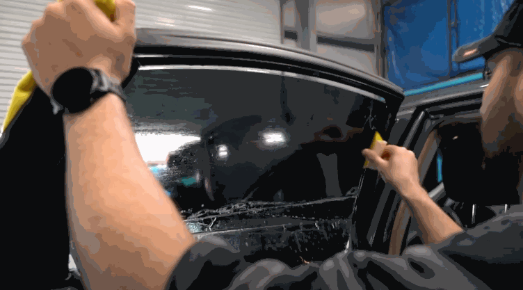 Installer uses a squeegee to apply ceramic window tint to a car window, removing water and air for a flawless finish. The darkened glass shows the enhanced privacy and sleek appearance achieved after installation.