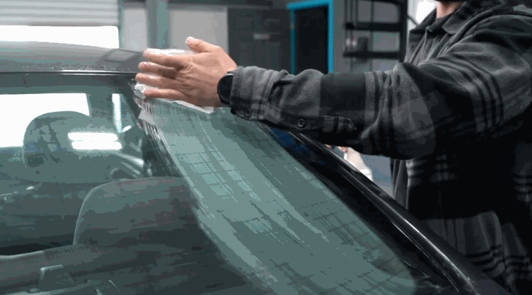 Technician carefully smoothing ceramic window tint film onto a car windshield inside a garage, ensuring a precise and bubble free application. The process highlights the hands on craftsmanship behind high quality tint installation.